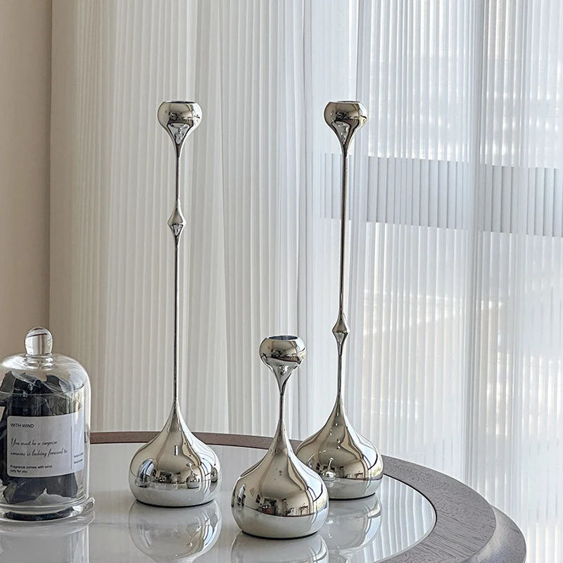 Bougeoirs décoratifs en métal argenté disposés sur une table ronde près d’une fenêtre.