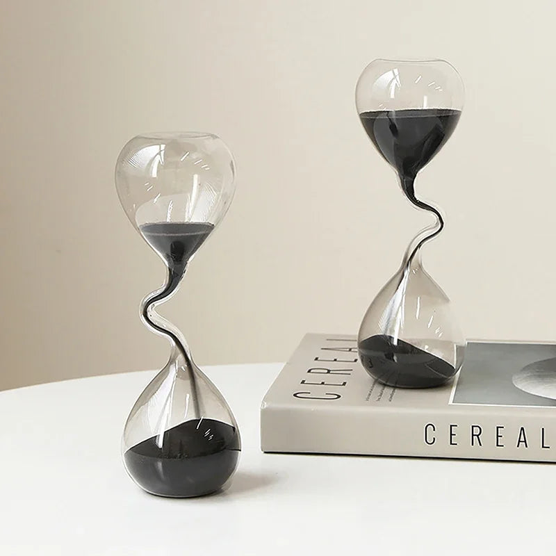 Sablier décoratif en verre avec sable noir, posé à côté de livres pour décoration intérieure.