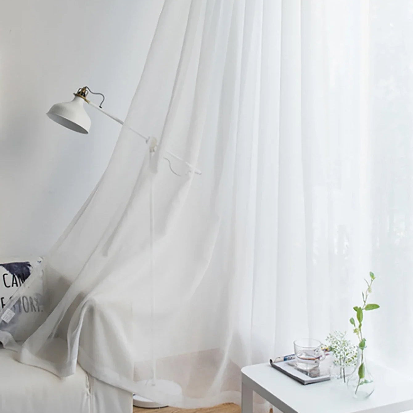 Rideau voilage blanc habillant une fenêtre de salon cosy, lampe liseuse et coussin en premier plan