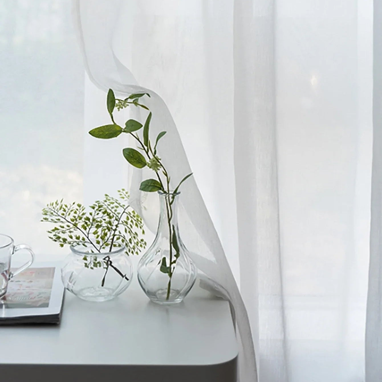 Voilage blanc translucide flottant devant une fenêtre, avec vases en verre et branchages sur console